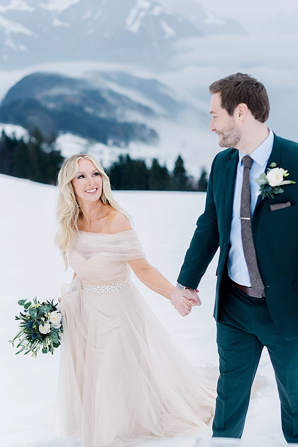 Couple portrait of bride and groom holding hands, walking in snow with white rose bouquet, pine trees and mountains behind them