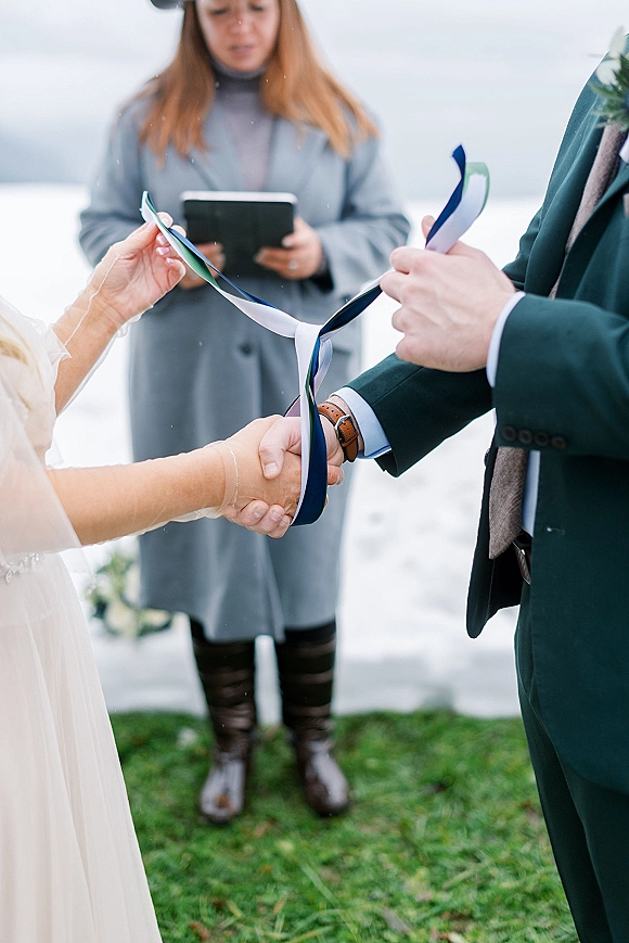 Wedding handfasting with a handfasting ceremony ribbon tying the bride and groom’s hands as an officiant holds a book on snowy grass lawn outdoors