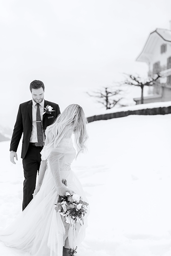 Couple portrait of a winter wedding couple in snow, bride in veil holding bouquet beside groom in suit under a pale winter sky