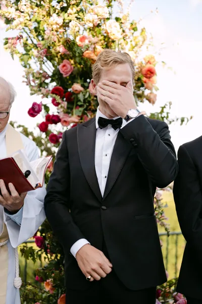 Ceremony moment with an emotional groom wiping tears in a black tuxedo under a rose and greenery floral arch on an outdoor lawn