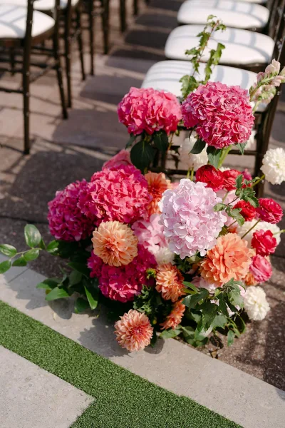 Ceremony aisle florals with hydrangeas, dahlias, and greenery arranged on the ground beside black chairs on an outdoor stone walkway