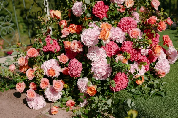 Wedding ceremony florals framing a wrought iron arch with pink and coral roses, hydrangeas, and greenery on a garden lawn by a stone patio