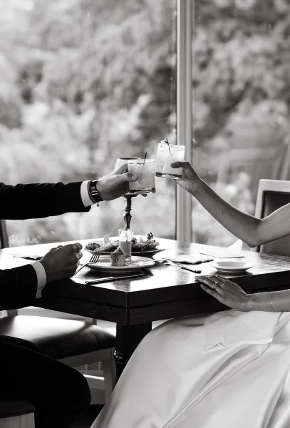 Wedding toast as bride and groom hands clink cocktail glasses, wedding rings visible over a dinner table by a window with trees outside