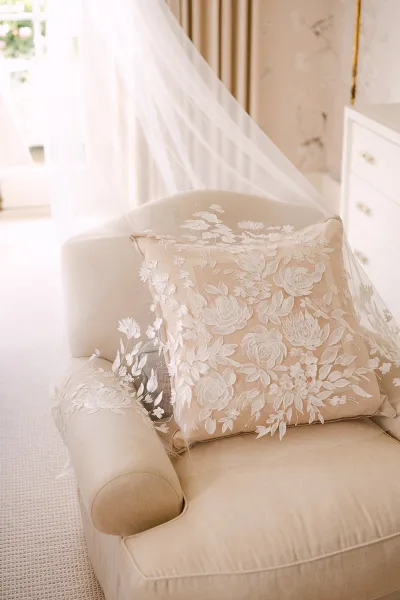 Wedding veil detail with floral lace applique draped over an embroidered pillow on an armchair, softly lit by window light in a neutral room