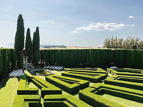 Outdoor ceremony setup with garden wedding ceremony rows of white chairs facing a statue and flowers by a reflecting pool in a hedge maze