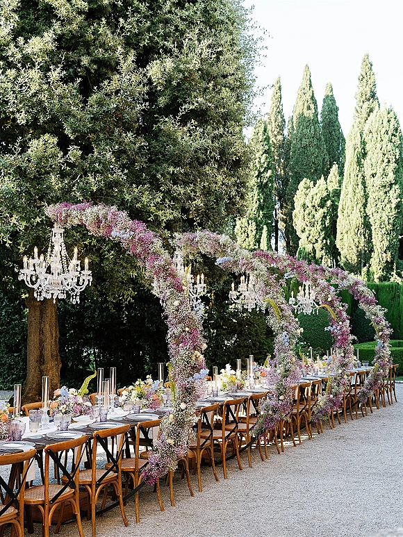 Outdoor reception tablescape for a garden wedding reception with long banquet table under floral arches and crystal chandeliers on a gravel path