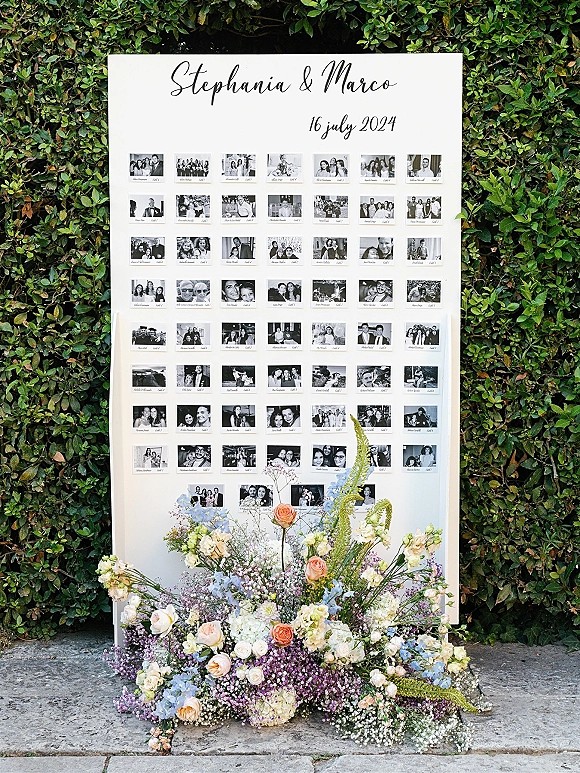 Wedding seating chart with photo escort cards and calligraphy header on an easel, accented by roses and hydrangea against a hedge wall