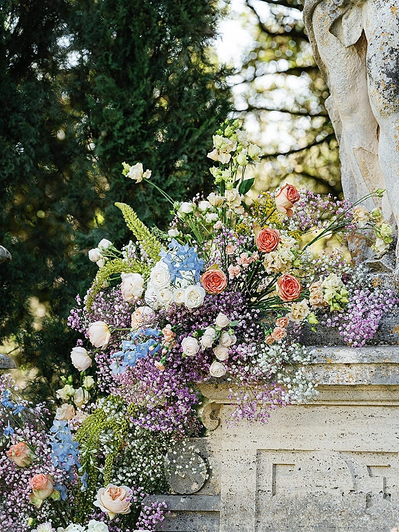 Wedding floral arrangement of roses, baby’s breath and delphinium in a stone urn on a balustrade in a garden with statue backdrop