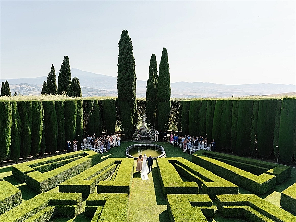 Outdoor wedding ceremony with white chairs lining a green hedge aisle, floral arrangements by a reflecting pool and fountain in a formal garden