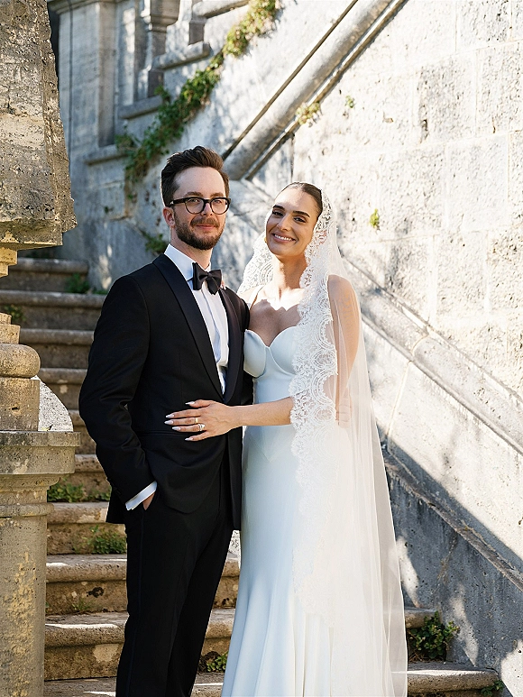 Couple portrait of bride and groom arm in arm, bride in lace mantilla veil and strapless dress, groom in black tuxedo on sunlit stone stairs