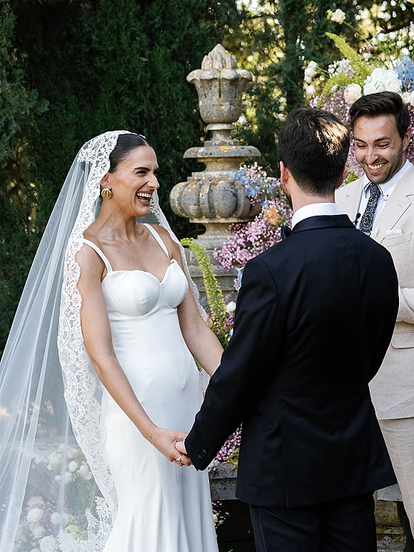 Wedding vows as bride and groom hold hands, bride laughing in strapless dress with lace veil at garden ceremony by stone urn fountain