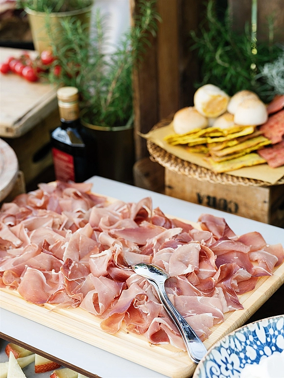 Charcuterie board with sliced prosciutto for wedding cocktail hour food, with crackers, bread rolls, serving tongs, and rosemary on a wooden table