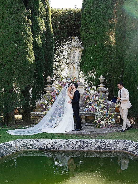 Ceremony kiss as bride in strapless gown and long lace veil kisses groom in tux on a stone terrace with garden florals by a reflecting pool