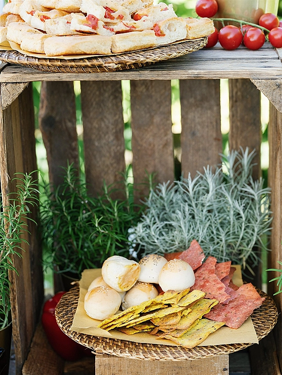 Wedding cocktail hour food on wicker trays and a rustic wooden crate stand with crackers, breadsticks, rolls, and herb garnish outdoors