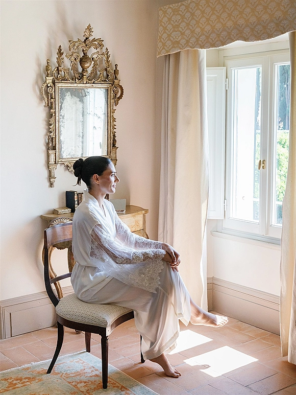 Bridal getting ready portrait of a bride in lace robe sitting by a window in a sunlit cream room, framed by an ornate gold mirror