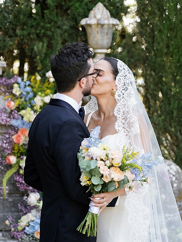 Wedding kiss portrait of bride and groom kissing, her lace veil and bouquet against garden greenery with stone urn and floral wall backdrop