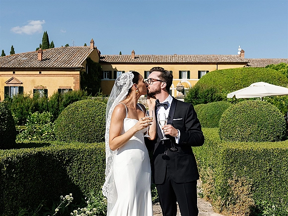 Wedding kiss as bride in lace veil and groom in black tuxedo clink champagne flutes on stone walkway by villa hedges under blue sky