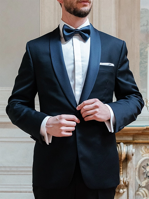Groom tuxedo with black tuxedo bow tie, satin shawl lapel and white pocket square, posed by ornate wall paneling and fireplace mantel