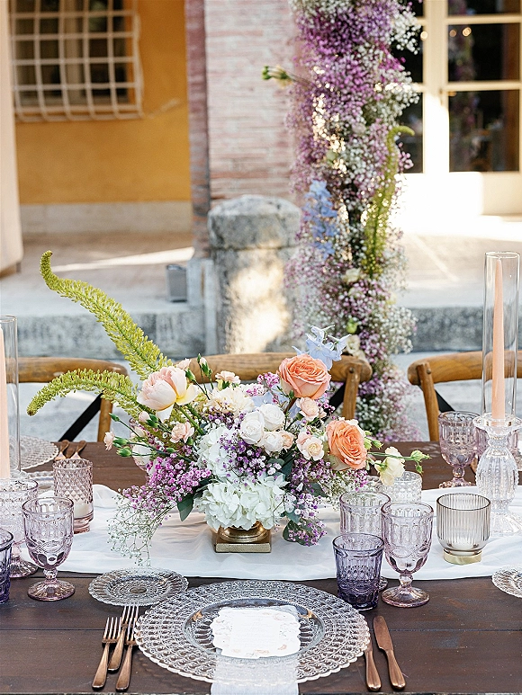 Reception tablescape with wedding table centerpiece of peach roses, hydrangea and taper candles, set on a wooden table in a brick courtyard