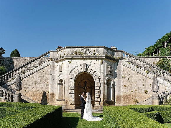 Couple portrait of bride and groom outdoors embracing full length, her veil trailing on a stone staircase by an arched doorway and fountain.
