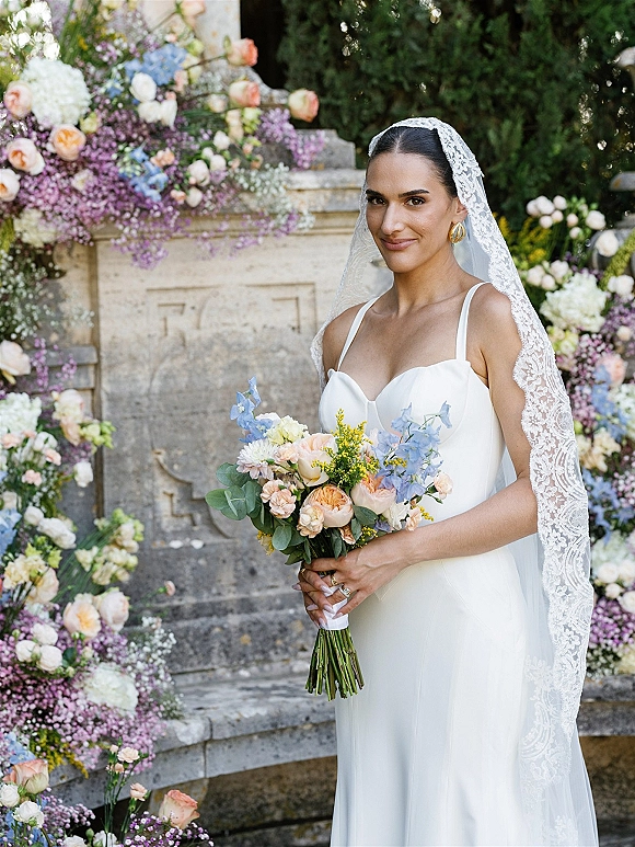 Bridal portrait of a bride holding bouquet with a lace veil, gold hoop earrings, and pastel florals by a stone fountain in lush greenery