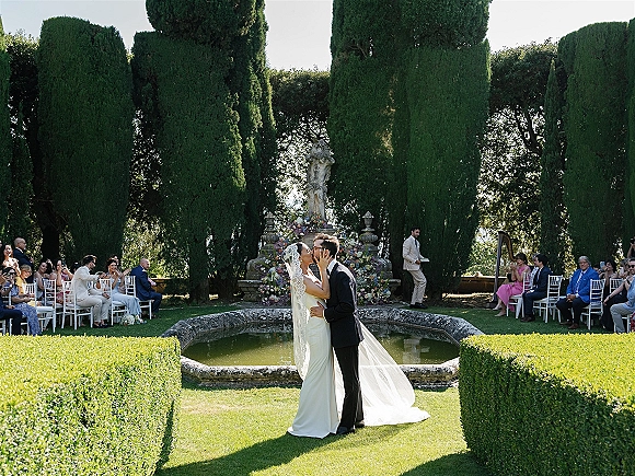 Wedding kiss as bride in strapless dress and veil kisses groom in black tux at garden ceremony, with fountain and seated guests behind