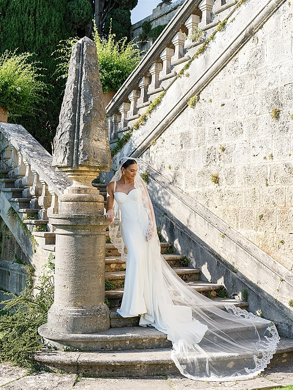 Bridal portrait of a bride in a strapless wedding dress with a lace-trim veil and long train on a sunlit stone staircase by greenery