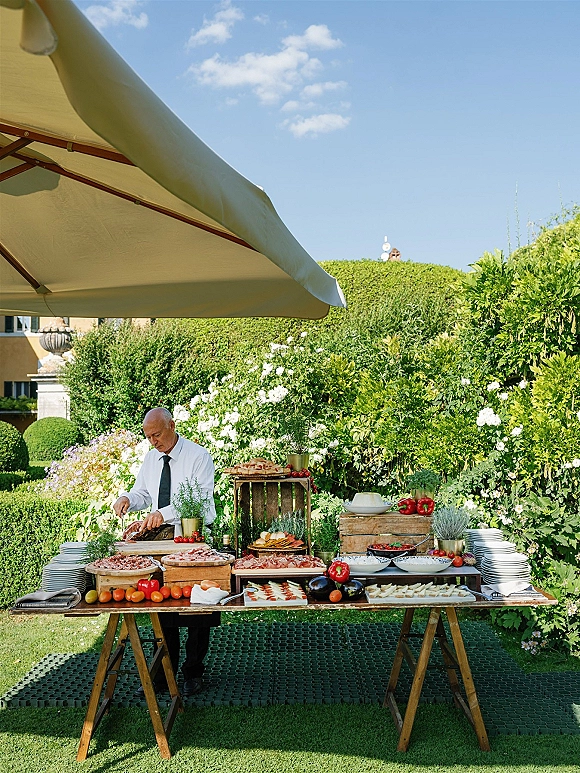 Wedding catering station outdoor wedding buffet with rustic wooden tables, charcuterie, cheese, bread, and potted herbs under an umbrella in a garden
