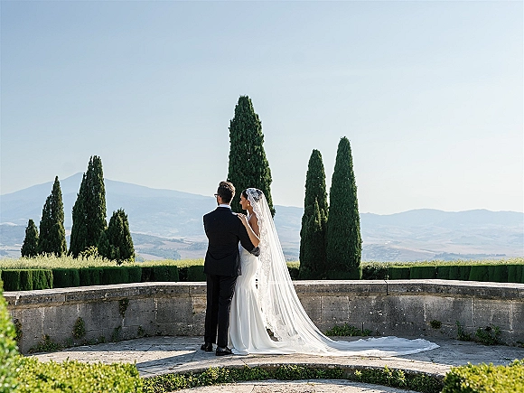 Couple portrait from behind, bride and groom overlooking mountain view with lace wedding veil trailing on a stone terrace by cypress trees