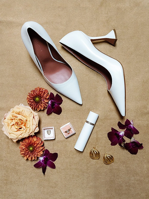 Bridal shoes flat lay with white pointed-toe heels, purple orchid accents, ring box, lipstick, and gold earrings on neutral paper backdrop