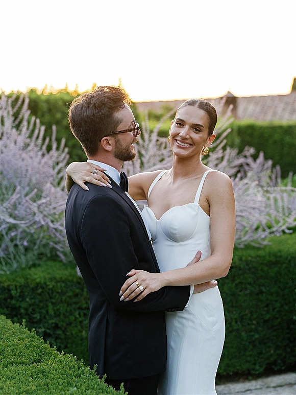 Couple portrait of bride and groom embrace, her white dress and his black tuxedo, in lavender hedges under a sunset sky