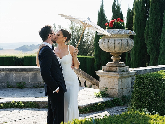 Wedding kiss as bride in strapless dress holds a white parasol, kissing groom in black tuxedo with glasses on a formal garden terrace