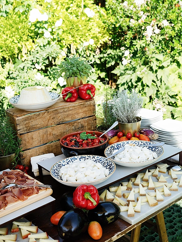 Wedding appetizer table with cheese wedges and prosciutto on platters and blue-and-white bowls, styled on wooden crates amid garden greenery