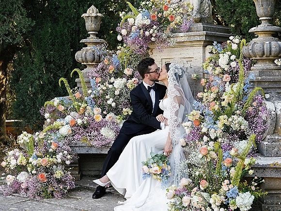 Wedding kiss portrait of bride and groom kissing on stone steps by a fountain, her lace veil and rose bouquet beneath a floral arch