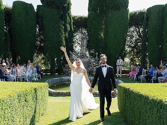 Wedding recessional as bride and groom walk the aisle holding hands, bride waving in veil beside guests in a garden with fountain statue