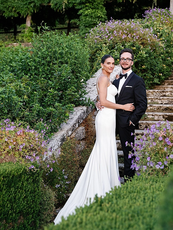 Couple portrait of bride in sleek gown and groom in black tuxedo hugging on stone steps, framed by lush greenery and purple flowers