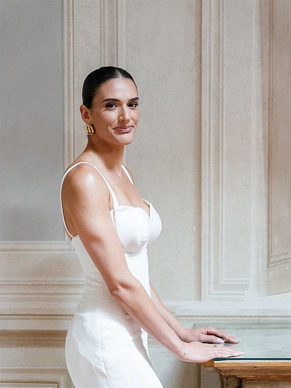 Bridal portrait of a bride in a minimalist bridal gown with spaghetti straps, gold hoop earrings, and ring against a paneled wall indoors