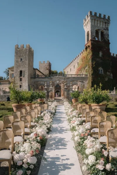 Outdoor ceremony setup along a garden ceremony aisle framed by terracotta planters, facing a castle stone facade under a clear blue sky.