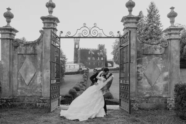 Wedding kiss portrait as the bride and groom dip beneath an iron gate and stone pillars, her veil trailing, with a castle and lawn beyond.