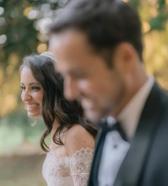 Couple portrait of a bride in lace gown with lace sleeves standing with her groom, smiling together in a sunlit garden beneath leafy trees.