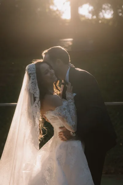 Wedding kiss portrait of groom kissing bride, her lace veil aglow, as they embrace in a sunlit garden with trees and lawn behind.