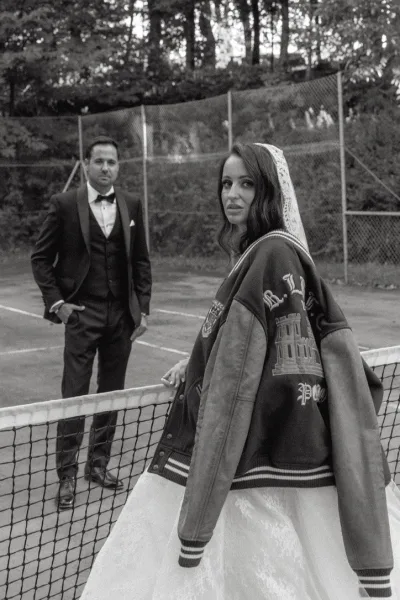 Couple portrait featuring a letterman jacket bride with her partner on a tennis court, bordered by a chain-link fence and trees.