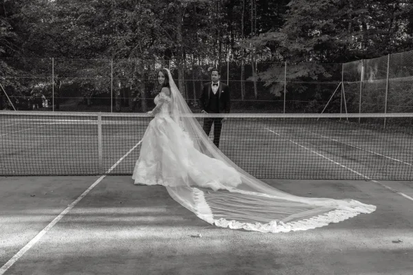 Couple portrait of a bride with long veil and a tuxedoed groom standing by a tennis net on a tennis court with chain-link fence and trees.