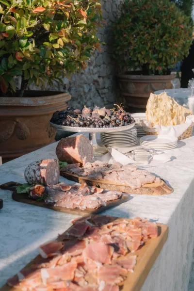 Charcuterie table for cocktail hour appetizers, layered with prosciutto, set in a garden beside potted plants and a rustic stone wall.
