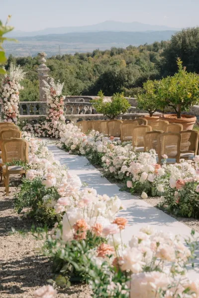 Ceremony aisle design featuring a flower lined aisle, framed by floral pillars on a stone terrace with garden trees and distant hills.