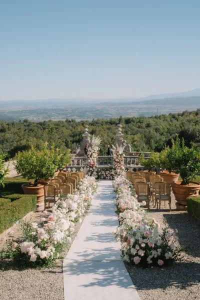 Ceremony setup showcasing outdoor ceremony decor with a white aisle runner on a stone terrace facing garden trees and distant hills.