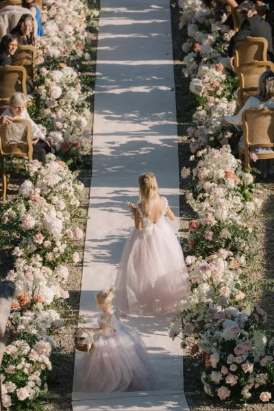Flower girl processional walking down the aisle, two little girls stride together, one clutching a flower basket along a gravel garden path.