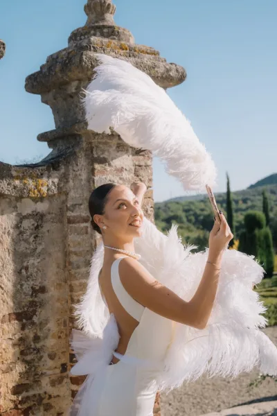 Bridal portrait of a bride with feather fan, in a sleek backless gown, by a stone wall with garden trees under a blue sky and distant hills.