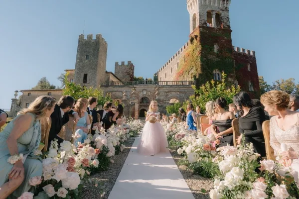 Flower girl processional makes a tender flower girl entrance, carrying a heart-shaped flower basket through a stone courtyard before a castle facade.