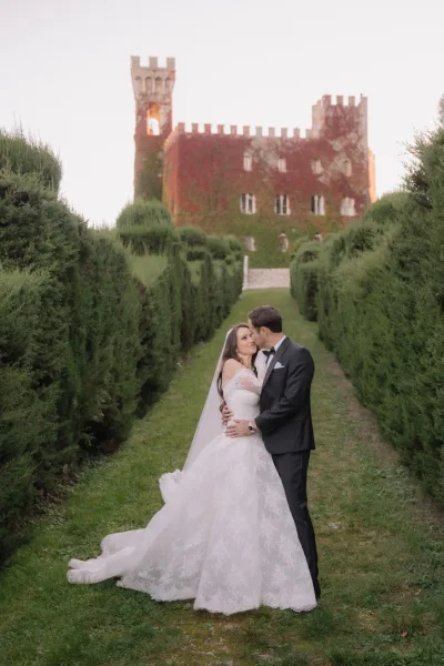 Couple portrait capturing a wedding kiss as the groom embraces the bride with a long veil before an ivy-covered castle and manicured hedges